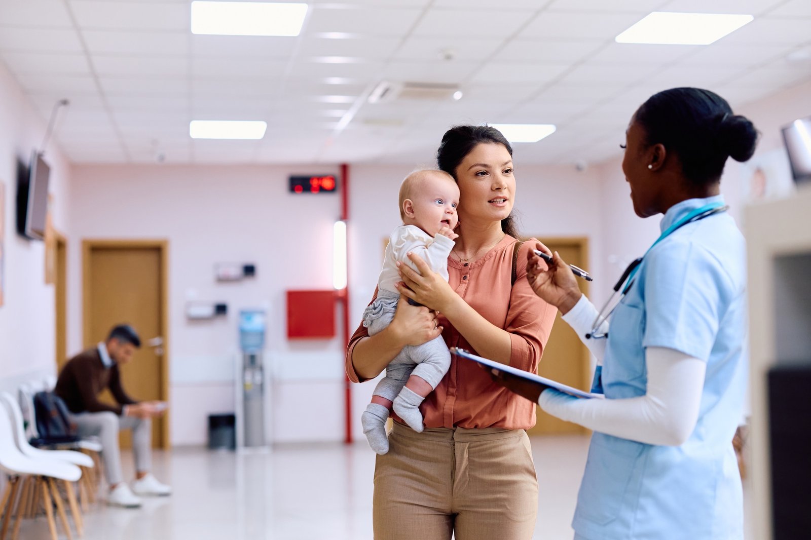 Mother with baby talking to nurse in hallway at pediatric clinic.
